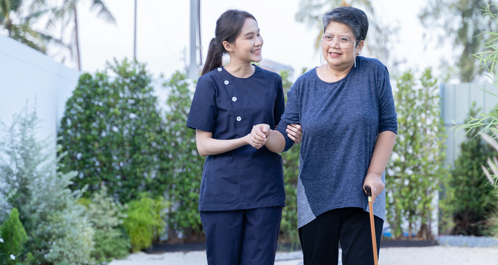 Woman walking with cane with a caregiver alongside her supporting her